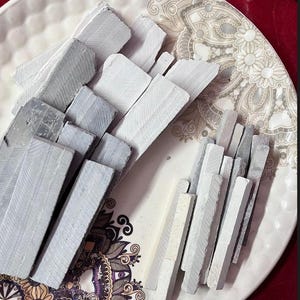 Op de afbeelding: Een verzameling grijze krijtstiften van verschillende formaten, gerangschikt op een decoratief wit bord met een bloemmotief. De krijtstukken hebben een rechthoekige vorm en zijn op elkaar gestapeld, wat een textuur geeft.