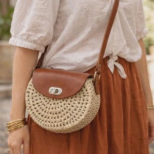 May include: A circular, woven crossbody bag with a brown leather flap and strap. The bag features a silver clasp and is worn with a white blouse and a rust-coloured skirt. Gold bangles are on the wrist.