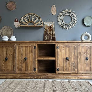 May include: A rustic wooden cabinet with four doors and a central open shelf. The cabinet has black metal ring pulls and hinges. Decorative wall hangings and a vase are displayed above the cabinet. A woven rug sits on the floor.