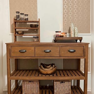 Puede incluir: Una isla de cocina de madera con tres cajones, un estante de listones y dos cestas de mimbre. Una especiero de madera y objetos decorativos se encuentran encima. La isla es de madera marrón claro con tiradores de cajón de metal.