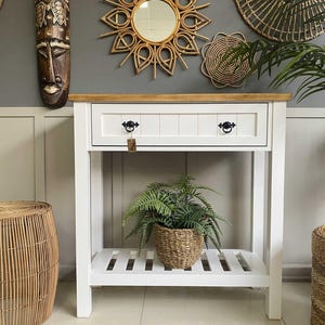 May include: A white ceramic mug with a black handle and the text "But First, Coffee" in a bold, black font. The mug is set against a blurred background, suggesting a cozy setting.