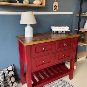 Könnte beinhalten: Eine rote Holzkonsolentisch mit vier Schubladen und Ringgriffen. Der Tisch hat eine natürliche Holzplatte und ein Lattenregal. Oben steht eine weiße Lampe mit beigem Schirm. Die Konsole steht in einem Raum mit blauen Wänden.