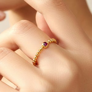 May include: A gold ring with a beaded band and a small, round, red gemstone. The ring is on a finger, and the image includes the brand name "SIMPLISH" in a stylized font.