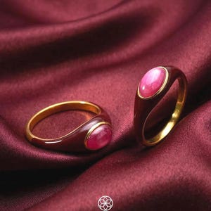 Op de afbeelding: Twee gouden ringen met ovale, roze edelstenen en donkerrode emaille zettingen worden gepresenteerd op een bordeauxrode satijnen ondergrond. De ringen hebben een klassiek design met een gepolijste afwerking. Het woord "SIMPLISH" staat in het wit onderaan.