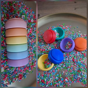 Può includere: Una composizione di oggetti rotondi dai colori vivaci, tra cui una pila di sei oggetti pastello. I colori vanno dal lavanda al rosa, arancione, giallo, turchese, blu e viola. Il tutto è posto su un letto di confettini colorati.