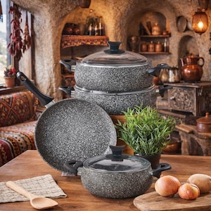 May include: A set of speckled gray cookware, including pots, pans, and lids, arranged on a wooden surface. The pots have black handles and glass lids. A wooden spoon and a small potted plant are also visible, suggesting a kitchen setting.