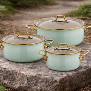 Può includere: Un set di tre pentole verde menta con bordi, manici e dettagli del coperchio dorati. Le pentole sono disposte su una superficie di pietra grigia, con uno sfondo naturale sfocato.