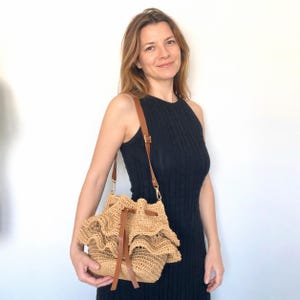 May include: A beige, woven shoulder bag with a drawstring closure and a brown leather strap. The bag has a ruffled design around the top and is held by a person wearing a black dress.
