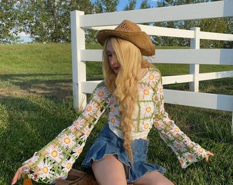 May include: A woman wearing a green and white crochet top with daisy flowers, a denim skirt, brown cowboy boots, and a brown cowboy hat. She is sitting in a grassy field with a white fence behind her.