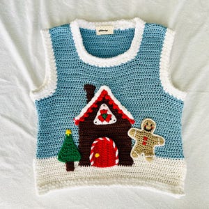 May include: A blue crocheted vest with a gingerbread house, a gingerbread man, and a Christmas tree design. The vest has a white trim around the neckline and armholes.