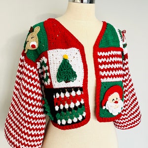 Puede incluir: Chaqueta de ganchillo hecha a mano en rojo, blanco y verde. La chaqueta con temática navideña presenta apliques de renos, árboles de Navidad y Papá Noel. Las mangas tienen un patrón de rayas rojas y blancas.