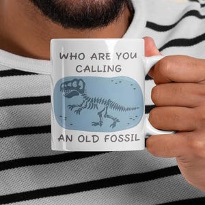 Puede incluir: Taza de cerámica blanca con el texto "WHO ARE YOU CALLING AN OLD FOSSIL" y un gráfico de esqueleto de dinosaurio azul. La taza es sostenida por una persona que lleva una camisa a rayas blancas y negras.