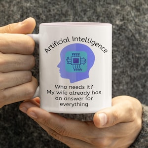 May include: White ceramic mug with a pink interior, featuring the text "Artificial Intelligence" and a blue graphic of a brain with a circuit board design. Below the graphic, the text reads, "Who needs it? My wife already has an answer for everything."