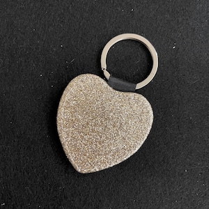 Puede incluir: Llavero en forma de corazón con purpurina dorada y un aro plateado. El corazón está cubierto de purpurina dorada y tiene un lazo de tela negra que lo conecta al aro. Fondo oscuro y texturizado.