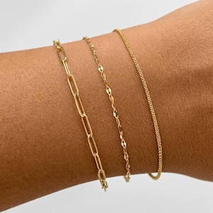 May include: Three gold bracelets of different styles are displayed on a wrist. The bracelets include a chain link, a delicate disc chain, and a thin, textured chain. The bracelets are all gold in color.