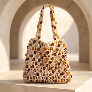 Puede incluir: Un bolso de ganchillo color crema con un patrón de rejilla adornado con cuentas translúcidas multicolores en tonos ámbar, marrón, verde y transparente. El bolso incluye dos asas de ganchillo a juego también decoradas con cuentas.