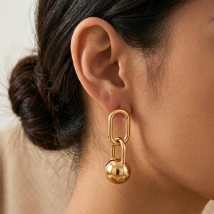 May include: Gold-toned earrings featuring a large oval shape connected to a smaller oval and a round, polished sphere. The earrings are shown on a person's ear, highlighting their reflective surface and elegant design. A modern jewelry piece.
