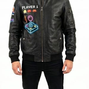 May include: Black leather bomber jacket with a zipper closure, ribbed cuffs, and a black collar. The jacket features a "PLAYER 1" graphic with retro video game icons and a joystick design. The person is wearing black pants and boots.