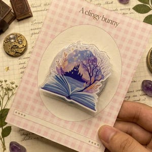 Könnte beinhalten: Ein klarer Aufkleber mit einem offenen Buch mit einer Burg- und Baumszene. Der Aufkleber befindet sich auf einer rosa-weiß karierten Karte mit dem Text "A clingy bunny". Weitere Artikel sind ein Kompass, kleine Bücher und Edelsteine.