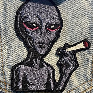 Op de afbeelding: Een geborduurde patch met een grijze alienfiguur met grote zwarte ogen omlijnd in roze. De alien is afgebeeld vanaf de borst en houdt een wit cilindervormig object met een rode punt vast, geplaatst op een lichtblauwe spijkerstof.