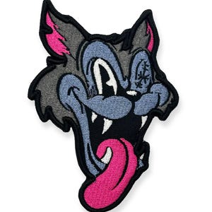 Puede incluir: Un parche bordado que muestra una cabeza de lobo de dibujos animados con pelaje gris, cara azul claro y una lengua rosa brillante que cuelga. El lobo tiene una amplia sonrisa con dientes visibles y orejas puntiagudas con detalles rosas.