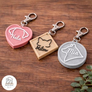 May include: Three pet ID tags with silver clasps. One is heart-shaped and pink with the name "Nala". Another is square and beige with a cat silhouette and the name "Milow". The third is round and grey with a dog silhouette and the name "Mia".