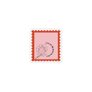 May include: A sticker with a pink background and a red postage stamp border. The design features a match lighting a flame with a heart in the center. The text "You Lit My Soul on Fire" is written around the flame.