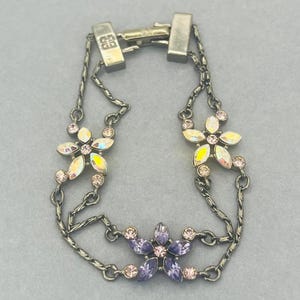 Puede incluir: Una pulsera plateada con tres diseños de flores. Las flores están adornadas con cristales iridiscentes y rosas. La pulsera tiene un diseño de doble cadena y un cierre de broche. La pulsera es una joya.