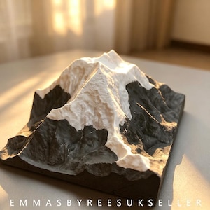 May include: A three-dimensional mountain model featuring a textured black base and a white snow-capped peak. The sculpture displays intricate topographical details and rugged slopes. The text EMMASBYREESUKSELLER is printed at the bottom.
