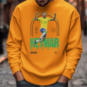 A man wearing a yellow sweatshirt with a graphic of a soccer player in mid-jump, celebrating.