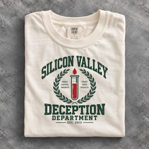 Op de afbeelding: Een gebroken wit T-shirt met ronde hals en een donkergroene opdruk met de tekst SILICON VALLEY DECEPTION DEPARTMENT EST. 2013. Het ontwerp bevat een reageerbuis gevuld met rode vloeistof, een rode druppel en lauwerkransen met de woorden IDEAS, CAPITAL, GROWTH, TRUST, DATA en RESULTS.