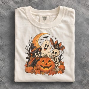 Op de afbeelding: Een gebroken wit t-shirt met een grafische print van een zwarte kat met een heksenhoed, een lachend spook, een skelet en een vleermuis rondom een grote pompoen. Het ontwerp bevat een maansikkel, herfstbladeren en kale bomen. Het neklabel vermeldt Comfort Colors 1717 L G.