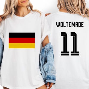 Puede incluir: Camiseta blanca con la bandera alemana en la parte delantera. La parte trasera de la camiseta muestra la palabra "WOLTEMADE" y el número "11" en negro. Una camiseta informal y unisex.