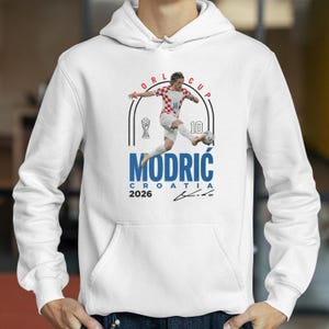 A white hoodie with a graphic design featuring a soccer player in action, along with text and a trophy icon.