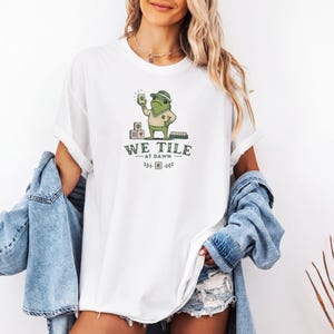Könnte beinhalten: Ein weißes T-Shirt mit einer Grafik eines grünen Frosches, der einen Hut und einen Pullover trägt und einen Mahjong-Stein hält. Der Text lautet WE TILE AT DAWN mit kleinen dekorativen Elementen. Das Shirt wird mit einer blauen Jeansjacke kombiniert.