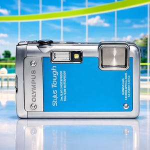 May include: Silver and blue Olympus Stylus Tough digital camera. The camera has a blue back panel with white text that reads "Stylus Tough" and "2m/6.6ft SHOCKPROOF 10m/33ft WATERPROOF". The camera has a flash and a zoom lens.