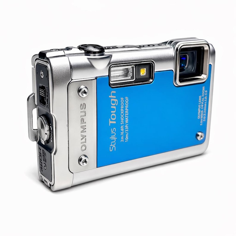 May include: Silver and blue Olympus Stylus Tough digital camera. The camera features a blue faceplate with the text "Stylus Tough" and "2m/6.6ft SHOCKPROOF 10m/33ft WATERPROOF". The camera has a lens and flash.