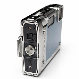 May include: A silver and blue digital camera with a rectangular shape. The camera has a silver metal frame and a blue back panel. It features a dial, buttons, and a small screen. The camera is designed for durability and outdoor use.