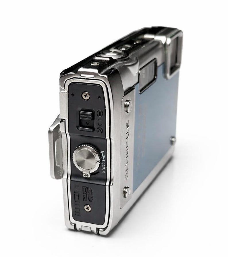 May include: A silver and blue digital camera with a rectangular shape. The camera has a silver metal frame and a blue back panel. It features a dial, buttons, and a small screen. The camera is designed for durability and outdoor use.
