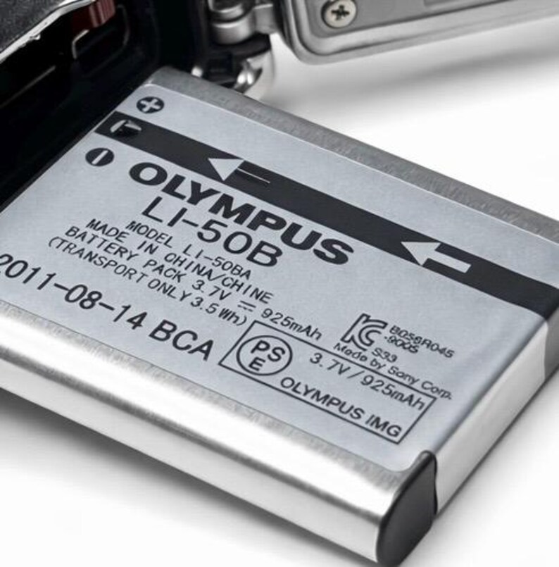 May include: A silver Olympus LI-50B camera battery, with black text indicating the model, voltage, and manufacturing details. The battery is partially inserted into a camera, with a metallic gray finish.