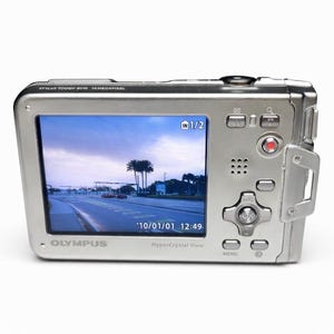 May include: A silver Olympus Stylus Tough-8010 digital camera with a screen displaying a road scene. The camera has multiple buttons and a red record button. The screen shows the date '10/01/01 12:49'.