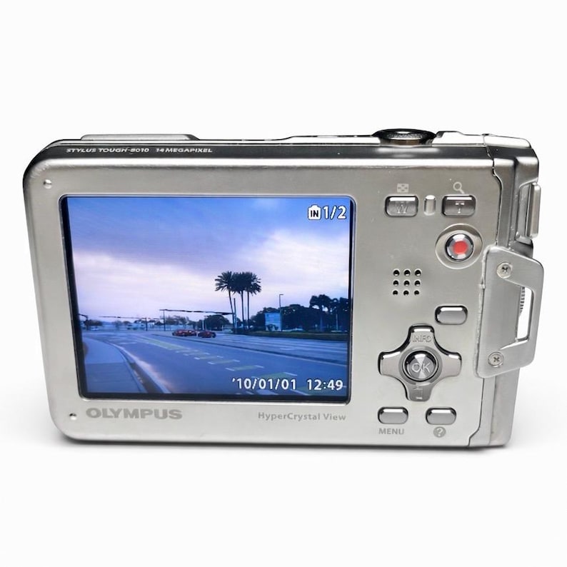 May include: A silver Olympus Stylus Tough-8010 digital camera with a screen displaying a road scene. The camera has multiple buttons and a red record button. The screen shows the date '10/01/01 12:49'.