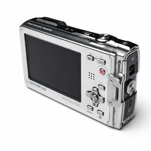 May include: Silver Olympus digital camera with a black screen and multiple buttons. The camera has a sleek, metallic design and is angled slightly to the right, showcasing its features. The Olympus logo is visible.