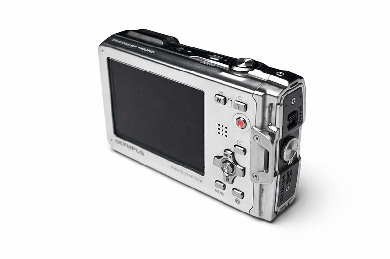May include: Silver Olympus digital camera with a black screen and multiple buttons. The camera has a sleek, metallic design and is angled slightly to the right, showcasing its features. The Olympus logo is visible.