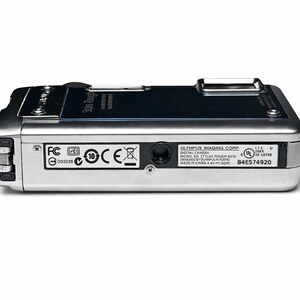 May include: Silver Olympus Stylus Tough-8010 digital camera. The camera has a black top panel and several regulatory and manufacturer labels on the bottom. The camera is designed in Tokyo and made in China.