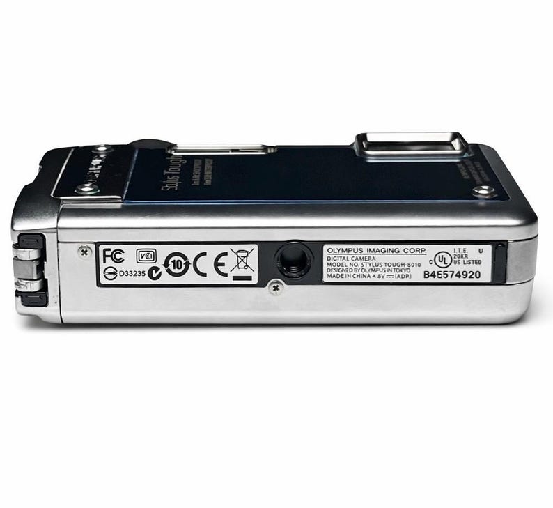 May include: Silver Olympus Stylus Tough-8010 digital camera. The camera has a black top panel and several regulatory and manufacturer labels on the bottom. The camera is designed in Tokyo and made in China.