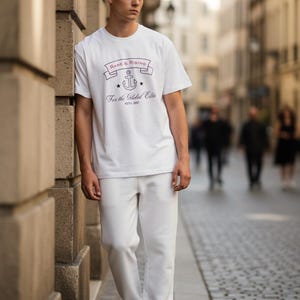 Pode incluir: Uma pessoa vestindo uma camiseta branca e cal&ccedil;as brancas. A camiseta tem um gr&aacute;fico com uma &acirc;ncora e o texto "Rare & Rising" e "For the Global Elite". A pessoa est&aacute; em uma rua de paralelep&iacute;pedos.