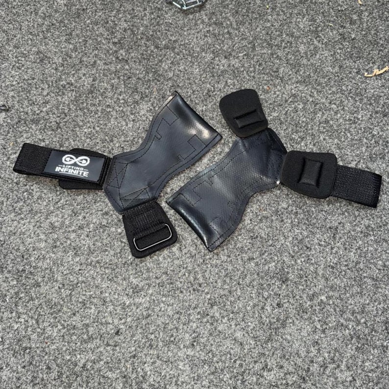 Puede incluir: Par de agarres de halterofilia negros con correas de mu&ntilde;eca ajustables. Los agarres est&aacute;n hechos de un material texturizado y presentan el texto "Lifting Infinite" con un s&iacute;mbolo de infinito.