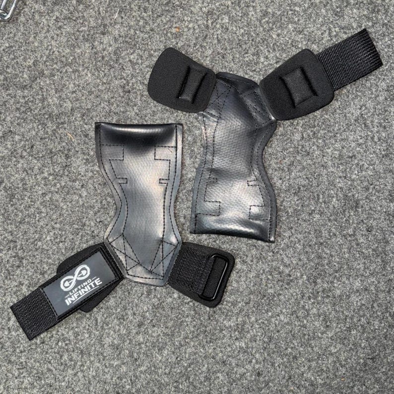 Puede incluir: Par de agarres de halterofilia negros con correas para la mu&ntilde;eca. Los agarres est&aacute;n hechos de un material duradero y presentan un dise&ntilde;o contorneado para un ajuste seguro. Las correas para la mu&ntilde;eca tienen el texto "Lifting Infinite" y un s&iacute;mbolo de infinito.