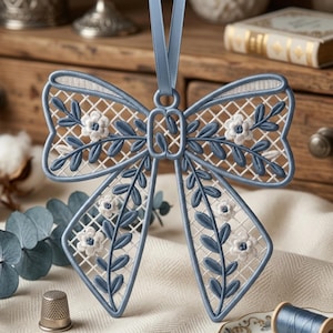 Peut inclure: Un ornement en forme de nœud décoratif avec une broderie complexe bleue et blanche sur une base en maille quadrillée. Le motif comprend des feuilles bleues brodées et de petites fleurs blanches à cœur bleu, suspendu par un ruban de satin bleu.